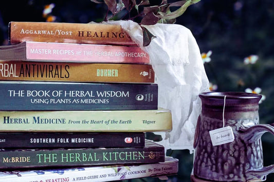 a stack of herbalism books next to a mug of herbal tea with a moody wildflower scene in the background