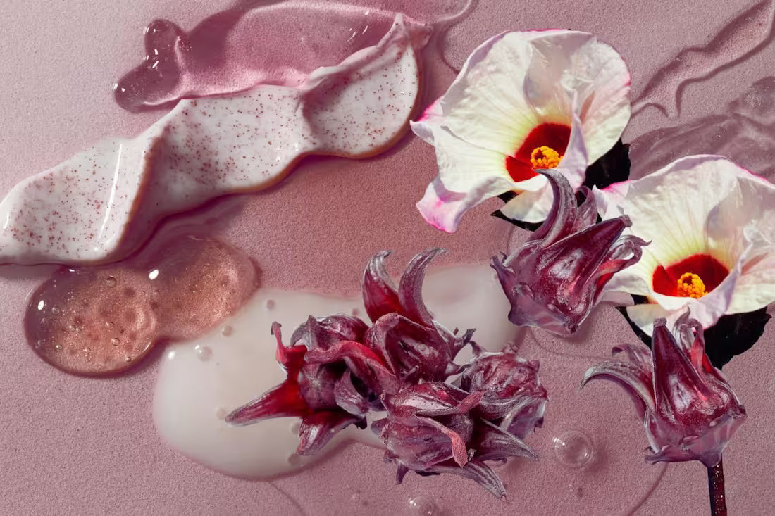 various pink-hued skincare liquids, lotions, and gels squirted onto a pink background with hibiscus calyces and white hibiscus flowers in the foreground