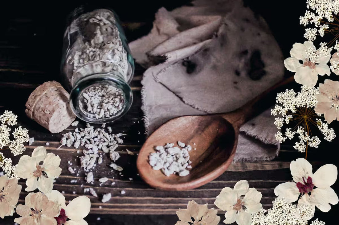 a glass jar filled with candied fennel seeds spilling out onto a dark wood surface with a wooden spoon and linen napkin and a decorative pressed flower overlay