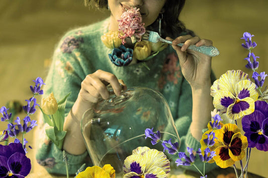 a woman holding a fork with fresh edible flowers on it, sitting at a table, holding a glass cloche surrounded by edible flowers
