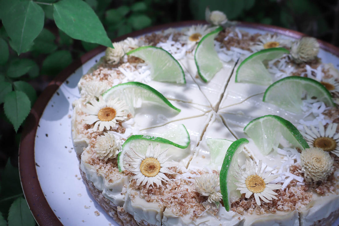 close up of a raw gluten free lime cheesecake sliced and decorated with limes and edible flowers on a ceramic plate with greenery in the background