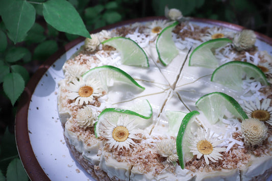 close up of a raw gluten free lime cheesecake sliced and decorated with limes and edible flowers on a ceramic plate with greenery in the background