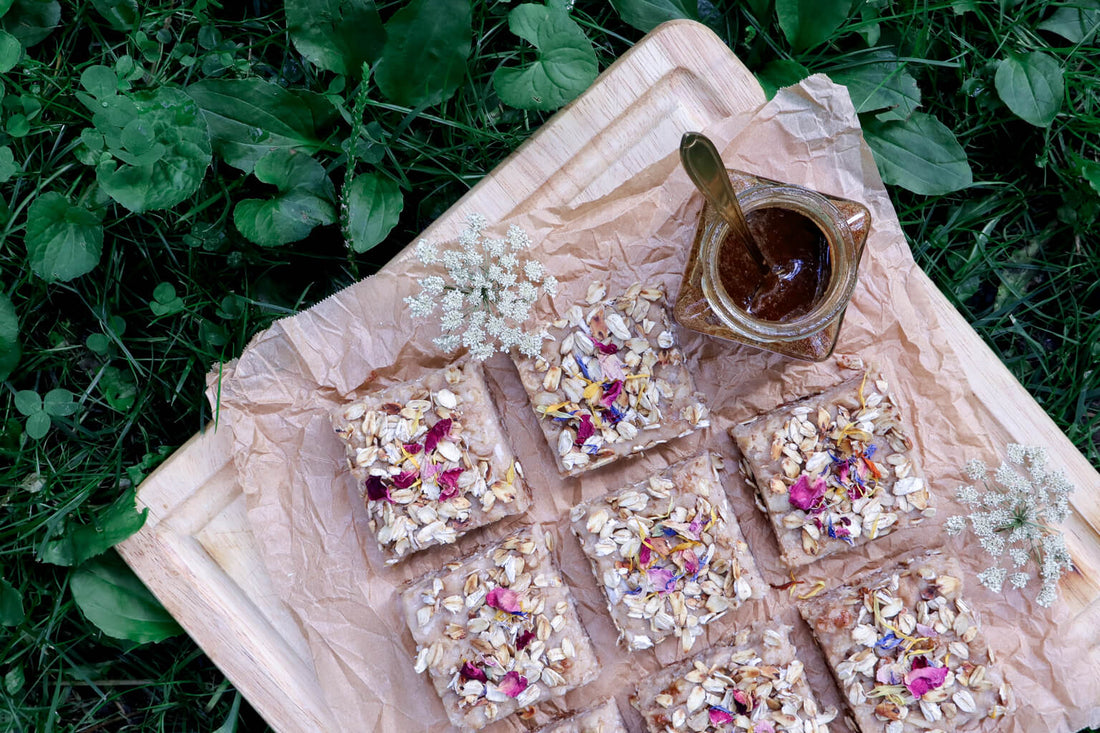 a cutting board with lemon oatmeal crumb bars decorated with dried edible rose petals and a jar of rose herbal honey against a greenery background