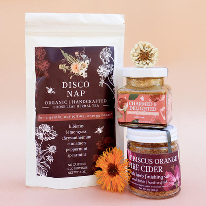 An herbal product bundle that includes a bag of looseleaf herbal tea, fire cider finishing salt, and a jar of rose herbal honey against a light pink background