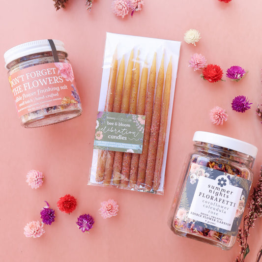 Three products on a pink background with flowers around them: a jar labeled 'Don't Forget the Flowers Edible Flower Finishing Salt', a package of beeswax birthday candles, and a jar labeled 'Summer Night FloraFetti Dried Edible Flower Garnish.