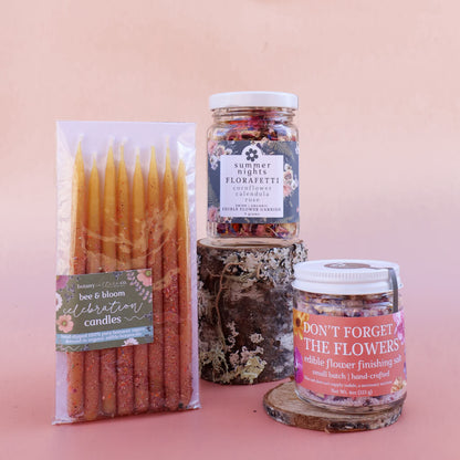 Three products on a pink background: beeswax candles, organic dried edible flower confetti, and a jar of floral finishing salt.