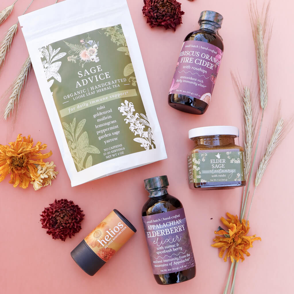 A collection of 5 Herbal products including a bag of loose-leaf herbal tea, bottles of elderberry syrup and fire cider, a jar of herbal honey, and a skin-soothing herbal salve on a pink background with dried flowers.