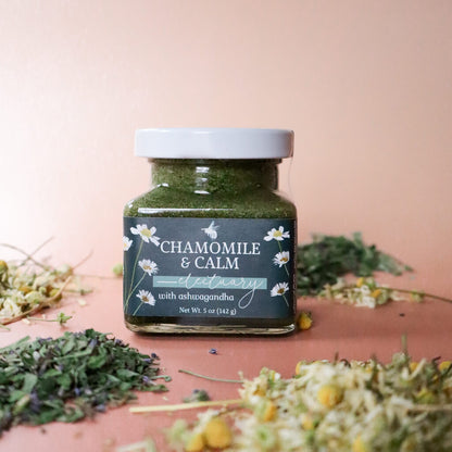 Jar of 'Chamomile & Calm Herbal Honey' product with dried herbs including chamomile and anise hyssop, on a pink background