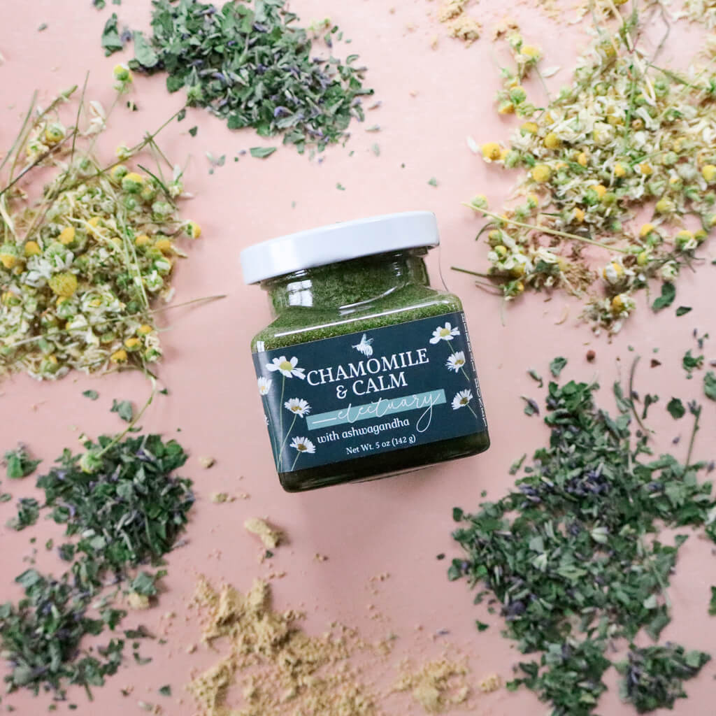 Jar of 'Chamomile & Calm Herbal Honey' surrounded by dried herbs including chamomile, anise hyssop, and ginger on a pink background