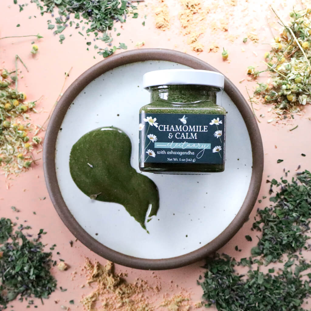 Jar of 'Chamomile & Calm Herbal Honey' on a white plate with a spread of green-colored herbal honey, surrounded by dried herbs on a pink background.