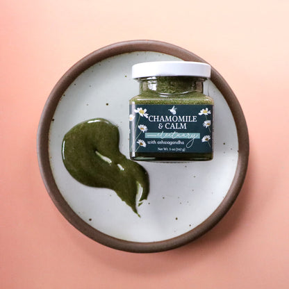 Jar of 'Chamomile & Calm Herbal Honey' next to a spread of green-tinted herbal honey on a white plate with a pink background