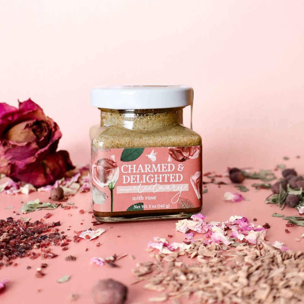 A jar of Charmed and Delighted herbal honey for heart and emotional support with visible floral and herbal ingredients, alongside scattered rose petals and other botanicals on a pink background.