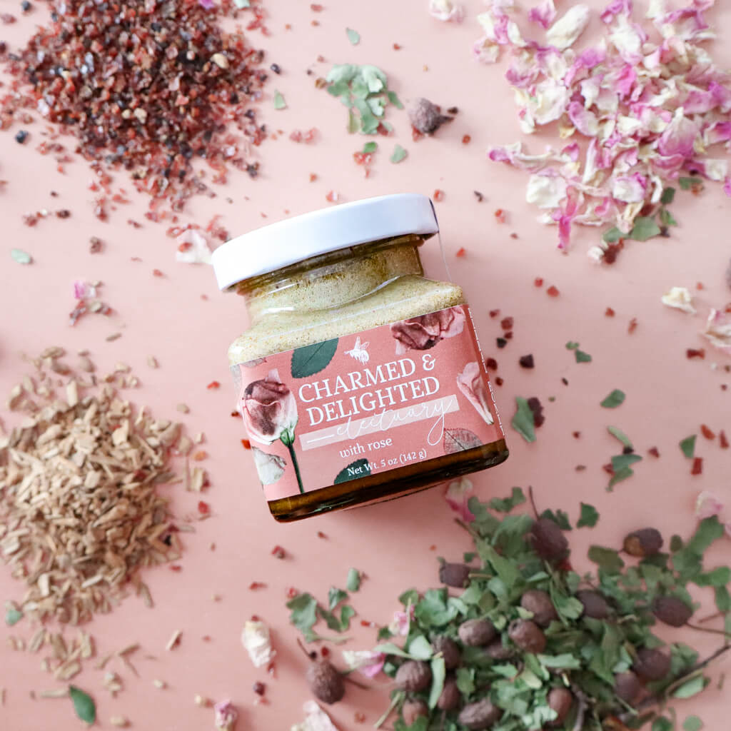 A jar of Charmed and Delighted herbal honey for heart and emotional support with visible floral and herbal ingredients, alongside scattered rose petals and other botanicals on a pink background.