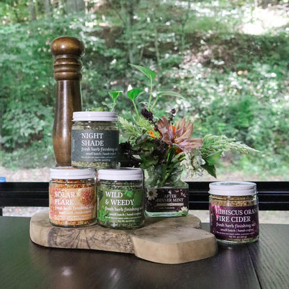 Collection of jars of herbal salts on a wooden dinning table in front of a window with a blurred green outdoor background