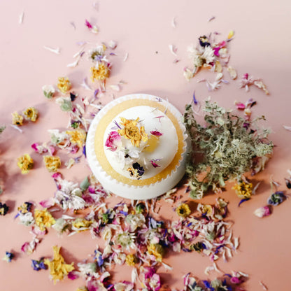 Aerial view of a vanilla cupcake with vanilla frosting decorated with dried edible flowers and surrounded by scattered dried edible flowers against a light pink background