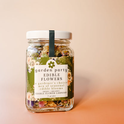A glass jar of Garden Party Edible Flowers with a green floral label that reads "A gardener's choice mix of seasonal edible blooms: dried organic edible flower garnish" against a peach gradient background