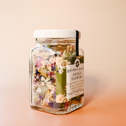 A side view of a glass jar of Garden Party Edible Flowers with a green floral label that reads "A gardener's choice mix of seasonal edible blooms: dried organic edible flower garnish" against a peach gradient background