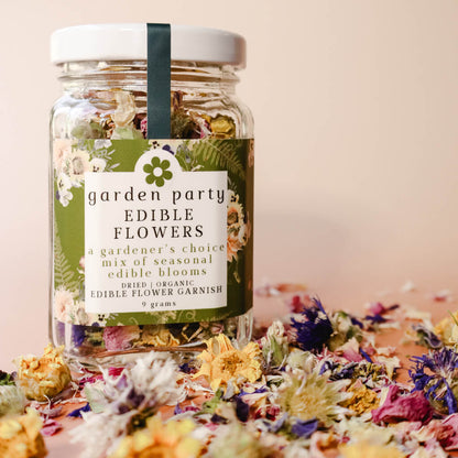 A glass jar labeled 'Garden Party Edible Flowers' with a variety of dried, colorful edible flowers displayed both inside the jar and scattered around it on a surface.