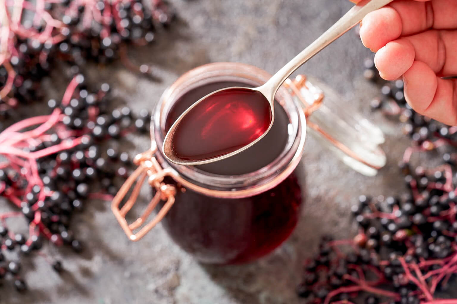 Jar of dark red elderberry syrup with a spoon, surrounded by elderberries on a stone surface