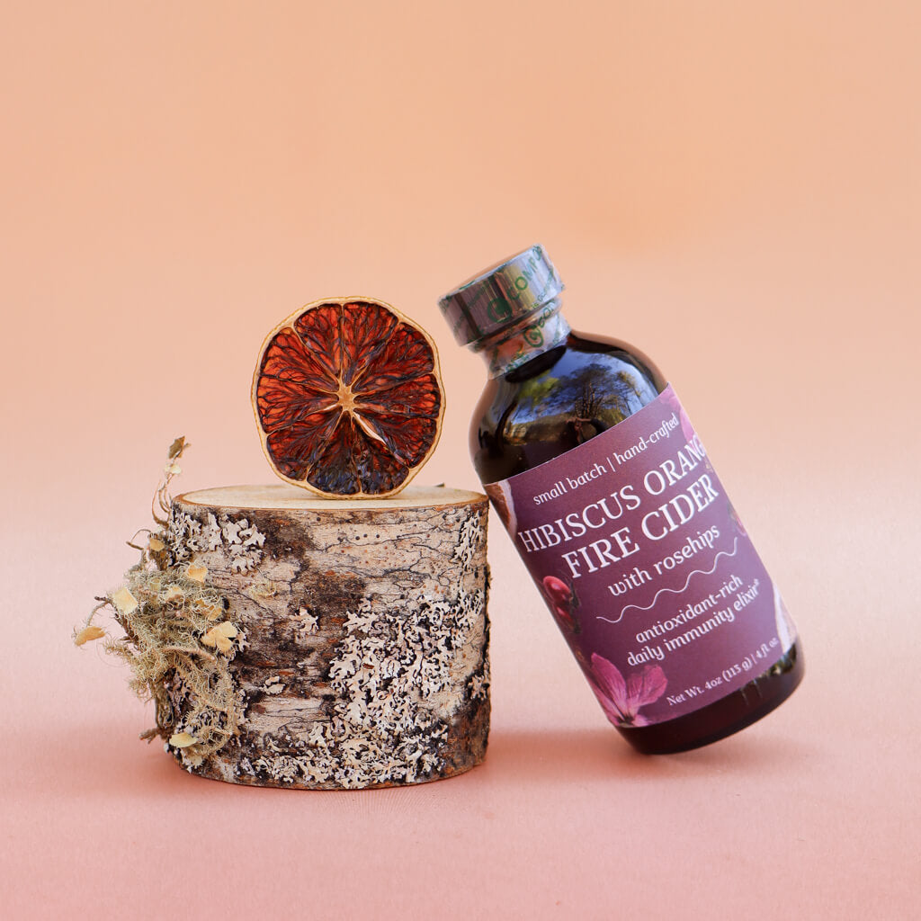 Bottle of Hibiscus Orange Fire Cider with a dried orange slice on a decorative wooden block against a pink background
