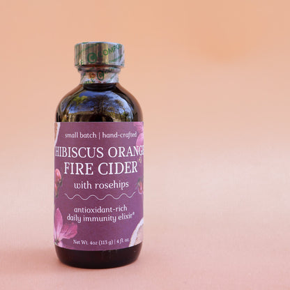 Bottle of Hibiscus Orange Fire Cider with rosehips for immune health on a pink background
