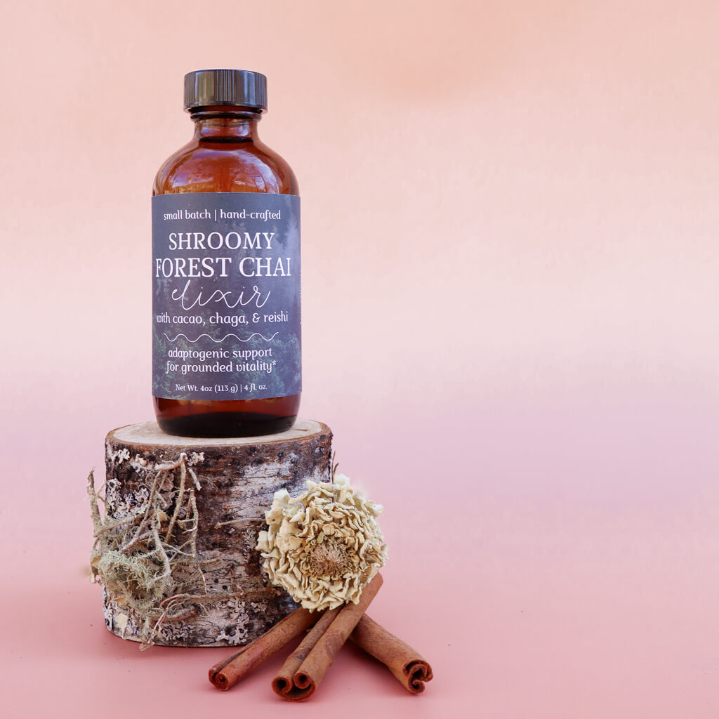 Bottle of Shroomy Forest Chai Elixir on a wooden block with cinnamon sticks and a dried herb on a pink background