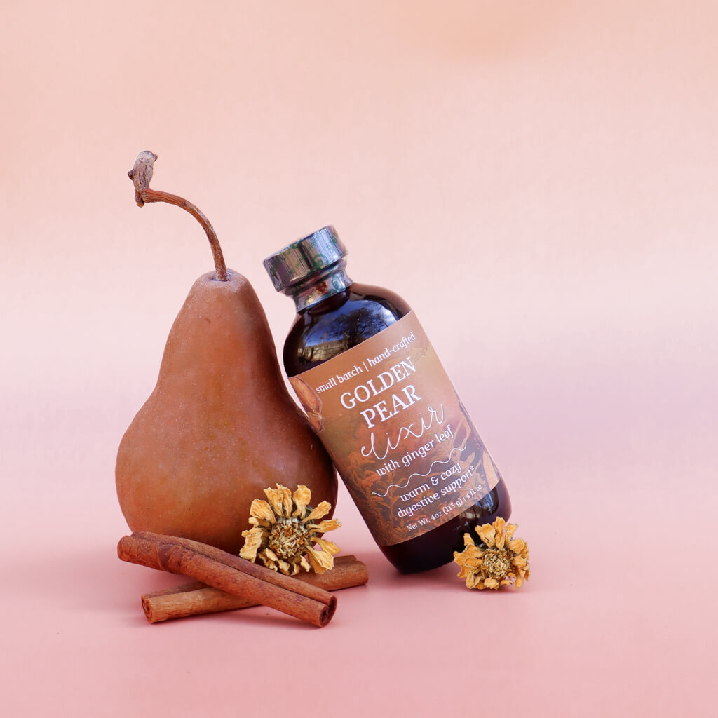Bottle of 'Golden Pear Elixir: Warming Digestive Support' with a pear, cinnamon sticks, and flowers on a pink background