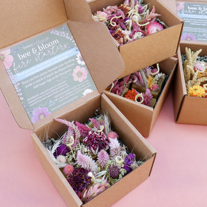 Open cardboard box set containing dried flower beeswax fire starters with a 'bee & bloom floral fire starters' label on a pink background