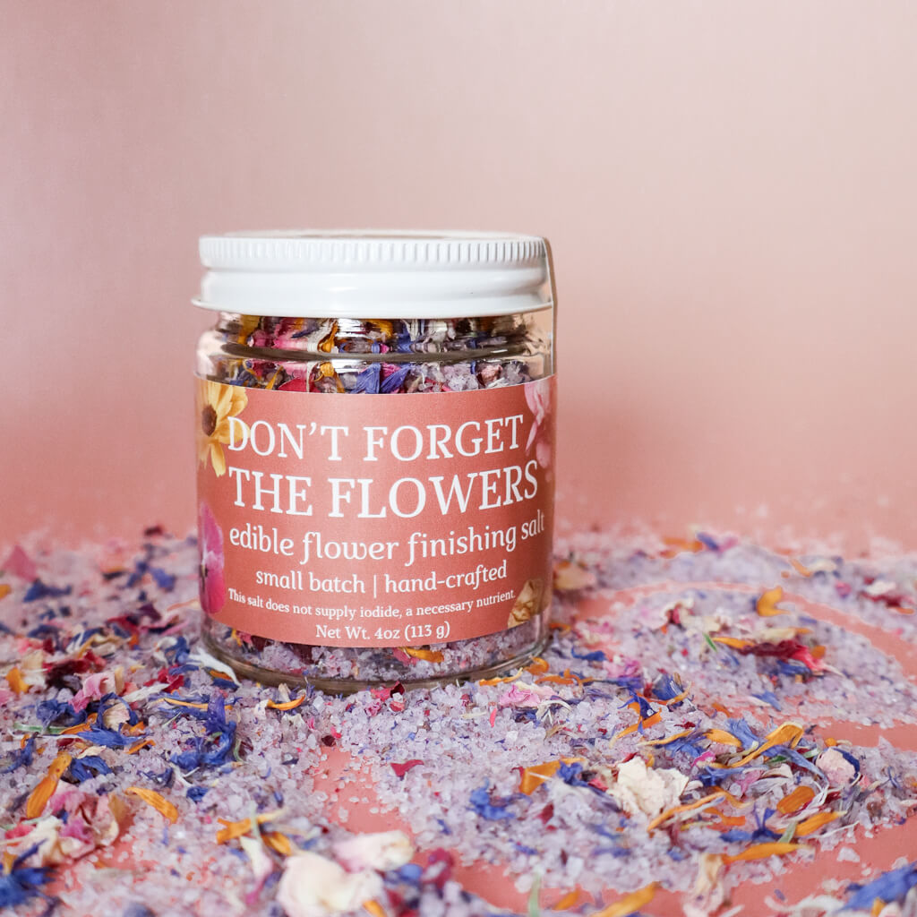 A jar of 'Don't Forget the Flowers' edible flower finishing salt, surrounded by scattered edible flowers in a variety of colors.