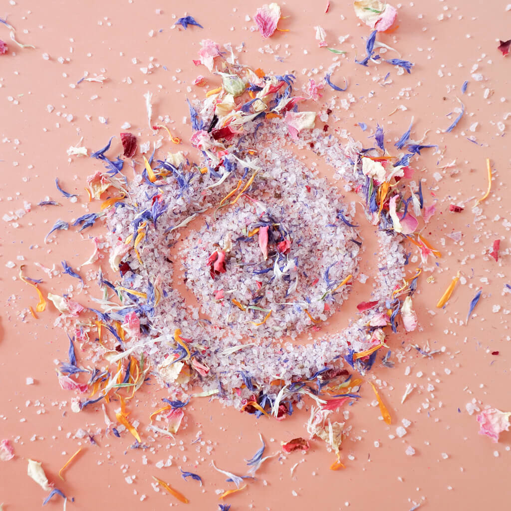 Aerial view of pile of 'Don't Forget the Flowers' edible flower finishing salt scattered on a light pink background. Coarse ground salt is a light lavender colors speckled with dried rose petals, blue cornflowers, and calendula petals.