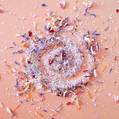 Aerial view of pile of 'Don't Forget the Flowers' edible flower finishing salt scattered on a light pink background. Coarse ground salt is a light lavender colors speckled with dried rose petals, blue cornflowers, and calendula petals.