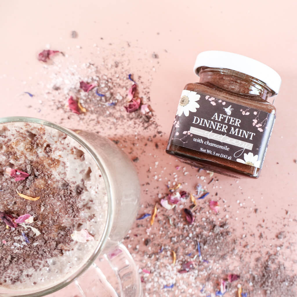 Jar of 'After Dinner Mint' herbal honey with a pink background, dried flowers, and a mug of iced cacao