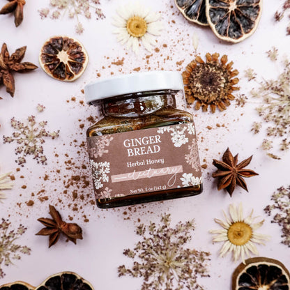 Jar of Gingerbread Herbal Honey surrounded by dried herbs and pressed flowers on a light background