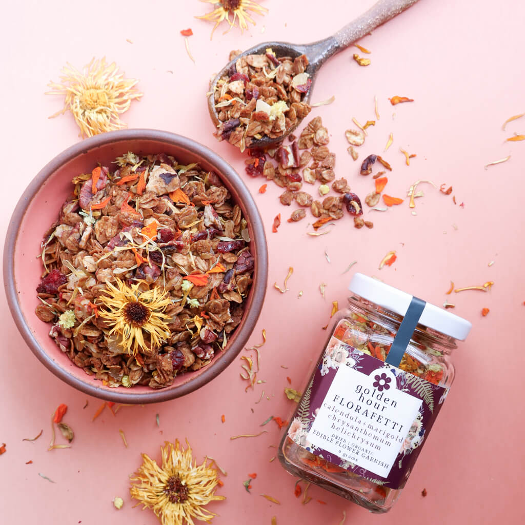 A pink bowl of gingerbread granola with edible calendula and marigold flowers with a clay spoon and a jar of "Golden Hour Florafetti Edible Flower Garnish" on a pink background with flowers.