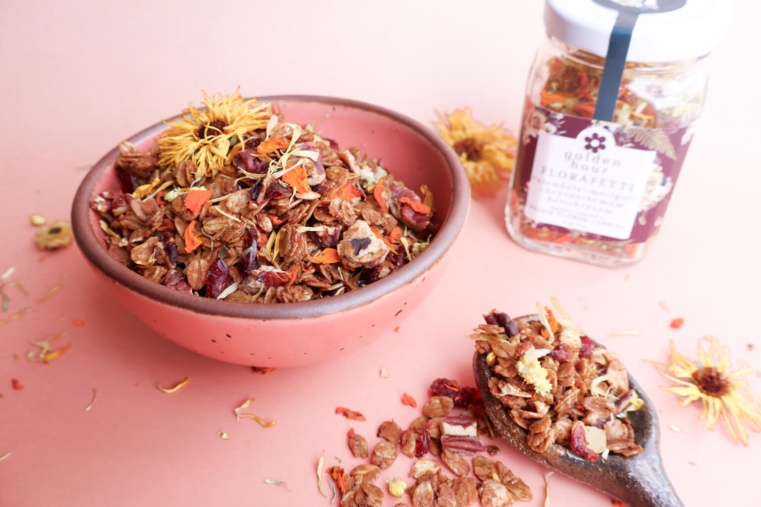 A pink bowl of gingerbread granola with edible calendula and marigold flowers with a clay spoon and a jar of "Golden Hour Florafetti Edible Flower Garnish" on a pink background with flowers.