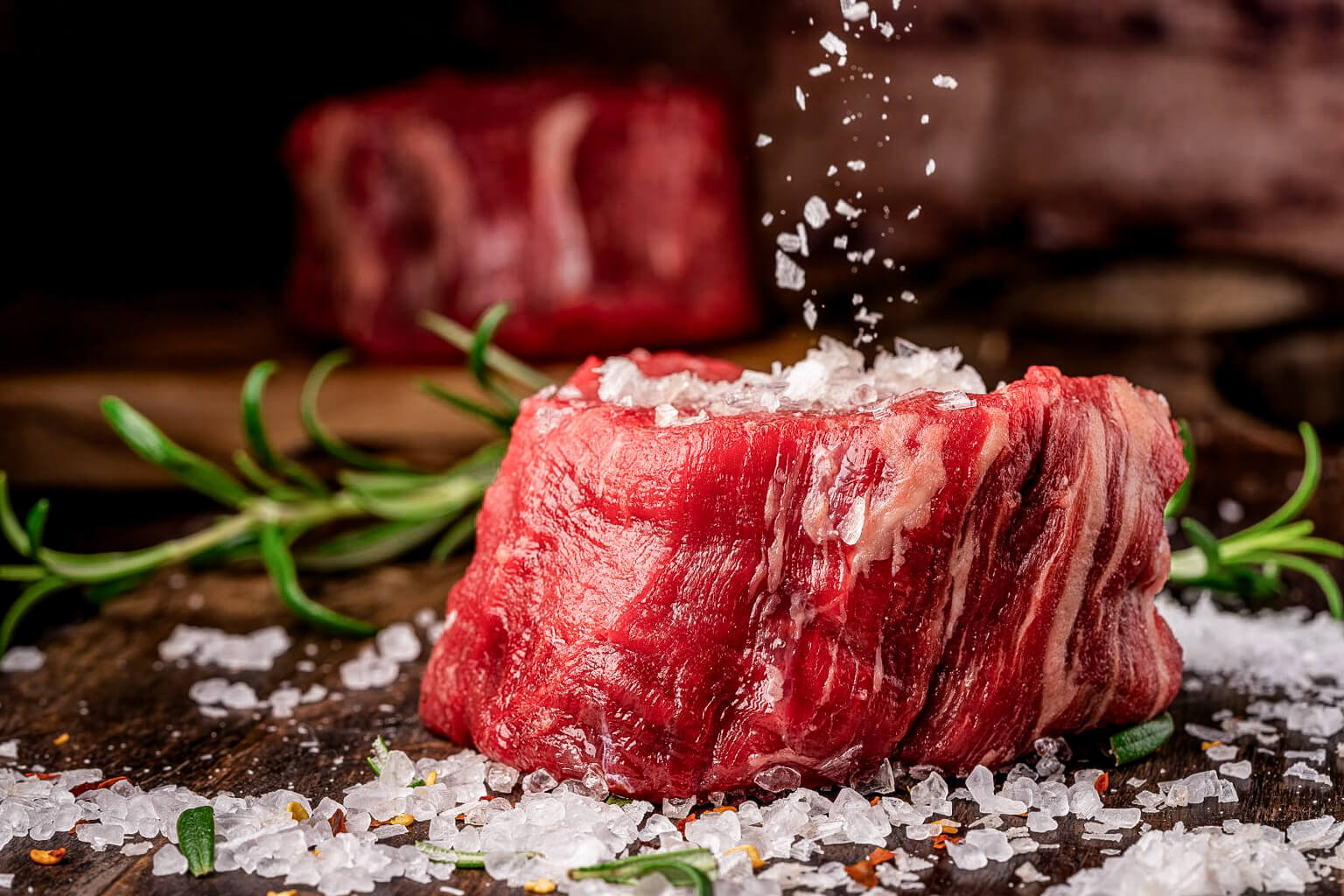 Raw steak being seasoned with salt on a wooden surface with rosemary.