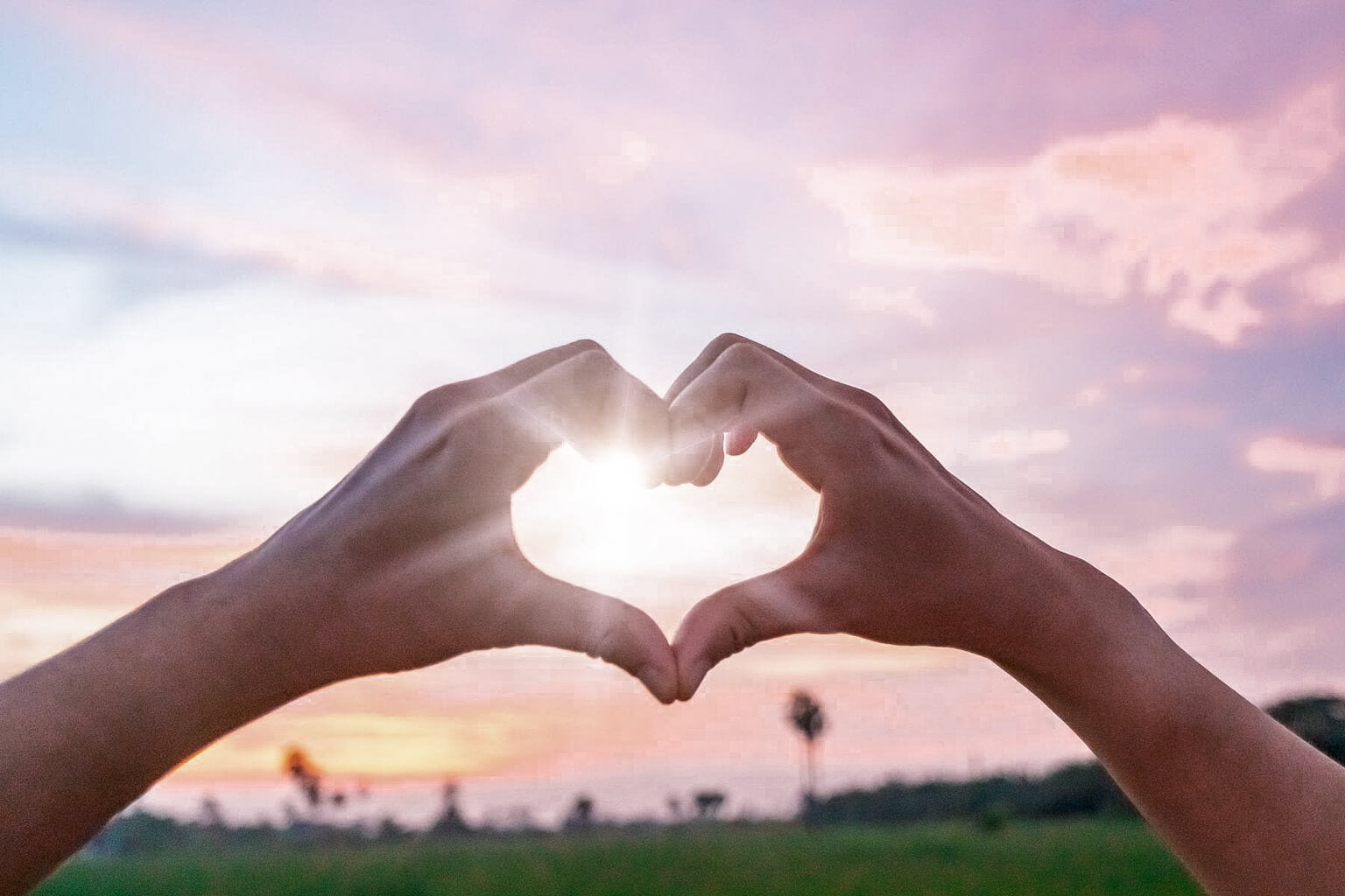 Two hands making a heart shape against a sunset sky.