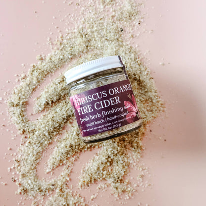 A glass jar of Hibiscus Orange Fire Cider finishing salt with citrus spice and herbs. Label shows the product name and a hint of orange color, placed on a surface scattered with loose coarse-ground sea salt.