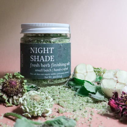 Jar of 'Night Shade' fresh herb finishing salt with herbs, garlic, and flowers on a pink background