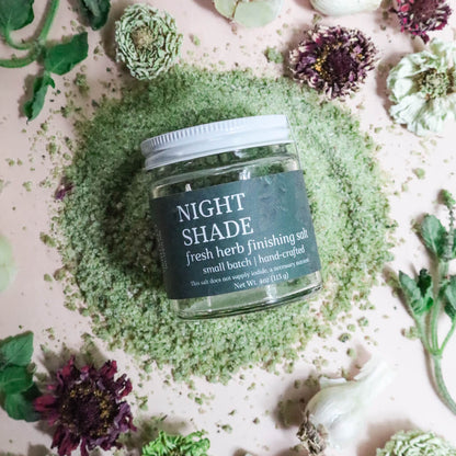 Jar of 'Night Shade' fresh herb finishing salt surrounded by green herbs and flowers on a light background