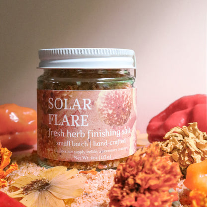 Jar of 'Solar Flare' fresh herb finishing salt surrounded by herbs and flowers on a neutral background