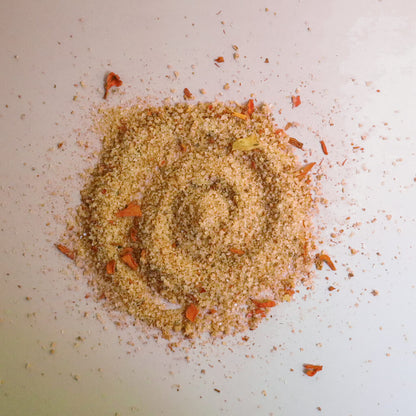 A pile of orange chili pepper herbal salt with flecks of dried marigold petals on a white surface