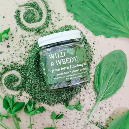 Jar of 'Wild & Weedy' fresh herb finishing salt surrounded by green-colored herbal mineral salt and fresh herbs like plantain leaves and chickweed on a light background