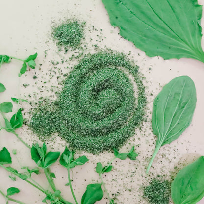 A pile of vibrant green Wild & Weedy Fresh Herbal Finishing Salt with fresh plantain leaves on a white background