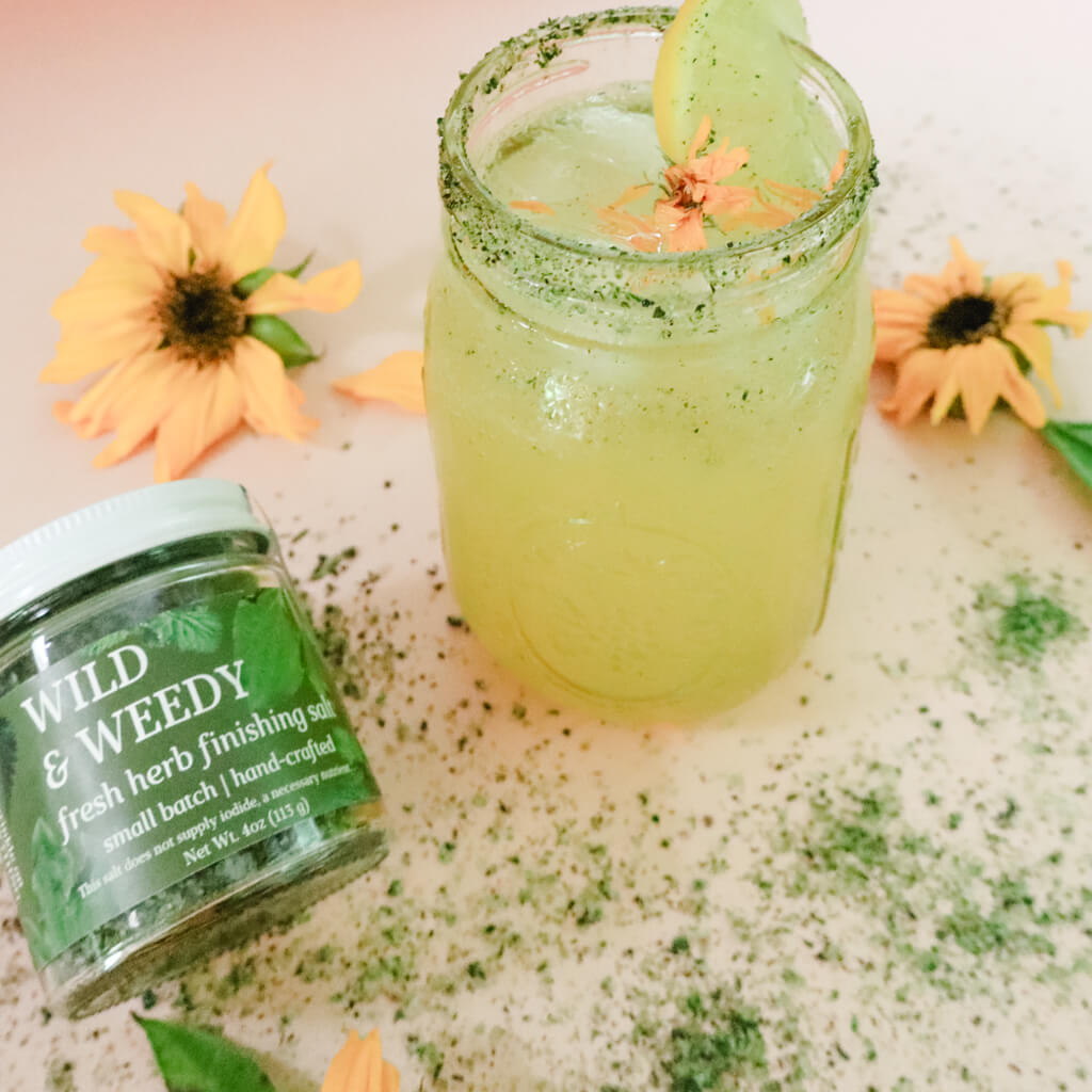 Glass jar of lemonade rimmed with green herbal salt next to a jar labeled 'Wild & Weedy Fresh Herb Finishing Salt' on a textured surface with flowers.