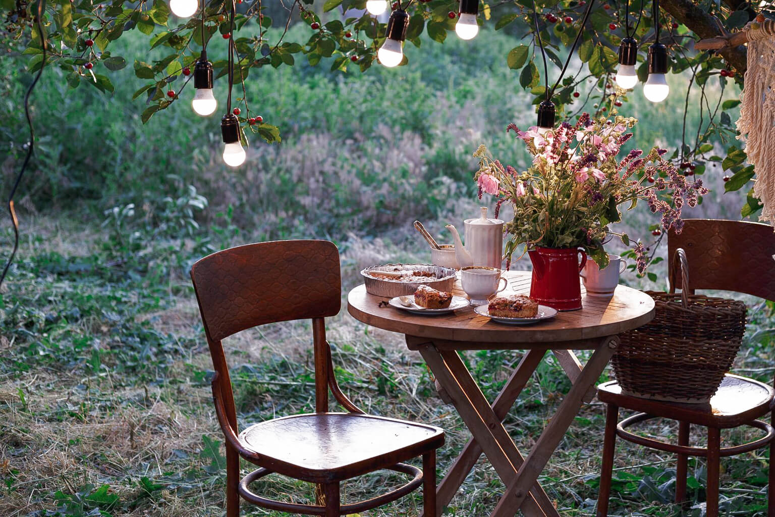 Outdoor garden party setting with a small table and chairs under string lights and flowers. Herbal tea pot and desserts decorated with edible flowers sit on the table.