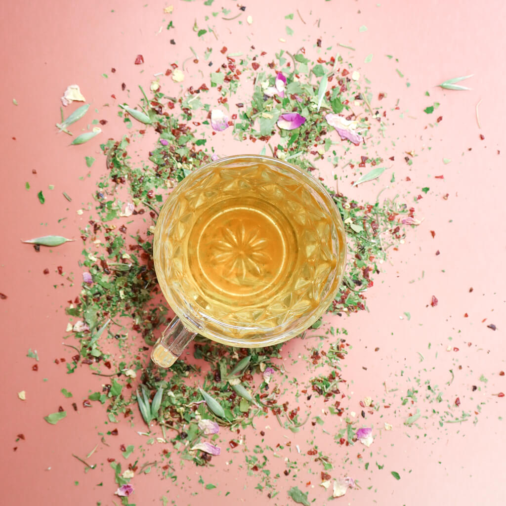 Aerial view of a vintage clear glass teacup filled with a light amber herbal tea against a light pink background and surrounded by loose herbal tea of pine rose petals, oat tops, rosehips, rosemary, and hawthorn leaf for heart health