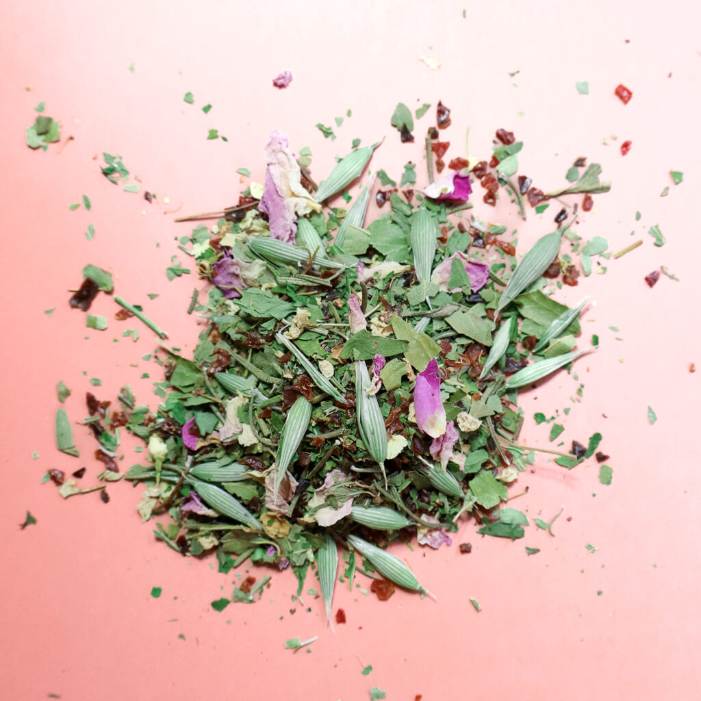 A pile of loose leaf tea made of hawthorn leaf, oat tops, rosemary, rose petals, rosehips against a light pink background