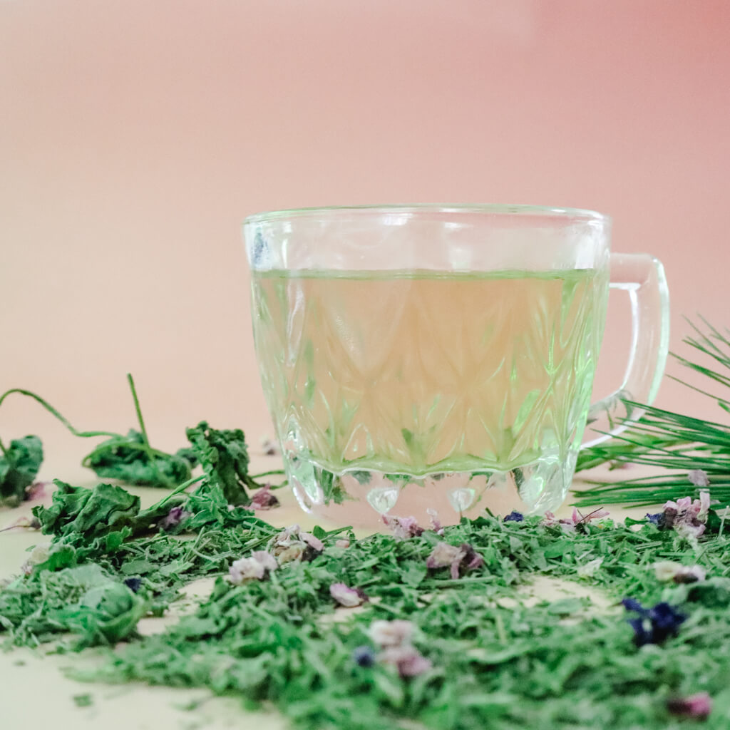 Clear glass mug filled with herbal tea surrounded by dried tea blend and flowers on a light pink background