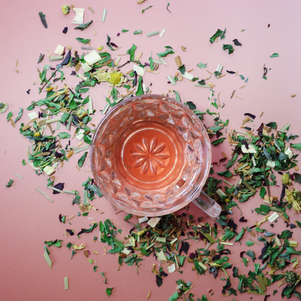 A cup of Disco Nap Herbal Tea – gentle energy support loose tea – herbal tea with various dried herbs and leaves scattered around on a pink background.